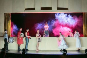 Männer und Frauen mit Instrumenten stehen vor einer Art Kasperletheater mit einer uniformierten Figur mit Pappmachékopf