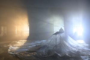 In a darkened, fogged room, with light coming in through three windows, there is a music stand under a large transparent film.