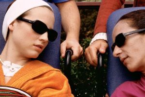 Ein Filmstill auf dem zwei weiblich gelesene Personen zu sehen sind, die eine Sonnenbrille tragen. Die linke Person trägt einen Verband um den Kopf und hat eine Trachealkanüle. Die Personen schauen sich an. Hinter ihnen sind zwei Personen, die die Rollstühle in denen die Frauen sitzen, schieben.