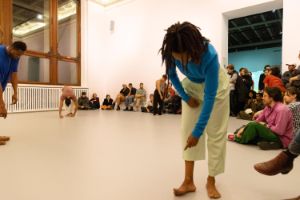 In einem hellrosa gestrichenen Raum findet eine Live-Performance mit dem Titel „study now steady“ statt. Mehrere Tänzer*innen bewegen sich dynamisch im Raum. Das Publikum beobachtet stehend und auf Bänken sitzend das Geschehen.