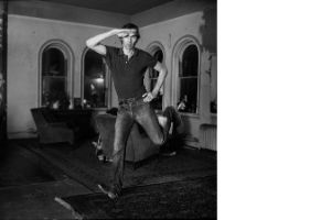 Eine Schwarz-Weiß-Aufnahme von einer Person in einem Raum mit vier Fenstern, Sesseln und Teppichen, die in die Luft springt und dabei eine Hand an die Hüfte und eine Hand an den Kopf hält.