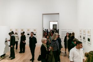 Besucher*innen in einem Ausstellungsraum. And den Wänden sind Rahmen mit Zetteln in Schwarz und Weiß.