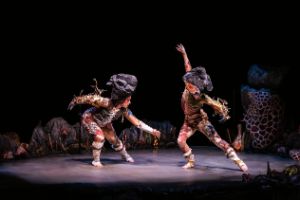 Two dancers wear stone-shaped headdresses and tight costumes with moss and bone-like elements.