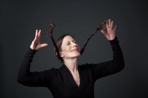 A woman in a black sweater throws her hands in the air, and her hair, braided into plaits, flies up too.