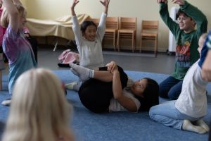 Kinder sitzen im Kreis und strecken ihre Arme in die Luft. In ihrer Mitte rollt sich ein weiteres Kind zu einem Päckchen zusammen.