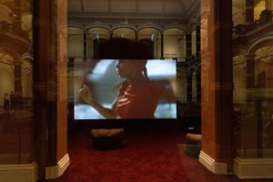 In einem Fenster, mit Blick auf den historischen Lichthof des Gropius Bau, spiegelt sich ein Bildschirm. Zu sehen ist eine Szene aus der Arbeit Arbeit „A Plot, A Scandal“ der der Künstlerin Ligia Lewis. Das angeschnittene Profil von Ligia Lewis in Bewegung ist schemenhaft zu erkennen.
