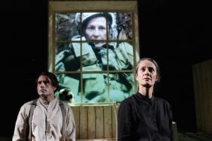 Two people look seriously straight ahead. Behind them in the window is a projection of a female soldier.