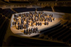 The musicians of an orchestra stand scattered across the stage of the Elbphilharmonie Hamburg with their instruments in hand.