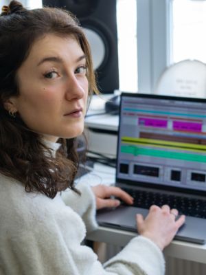 Eine junge Frau sitzt am Laptop und blickt über die Schulter direkt in die Kamera. Sie trägt einen hellen Pullover.