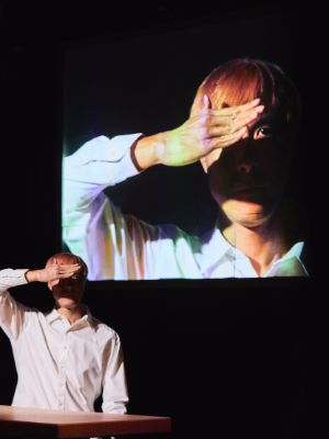 The artist wearing a white shirt stands at a high table and shields part of his face with his hand, so that one eye stands out brightly from the shadows. A projection in the background shows him up to his chest.