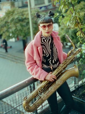 Sofia Salvo mit Saxofon und rosa Plüschjacke auf einem Balkon.