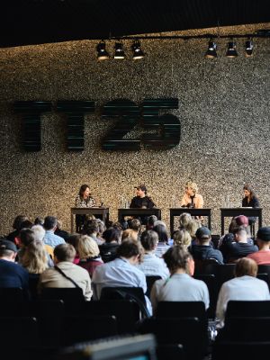 Four people are seated at tables lined up on stage, with the audience visible in the foreground.