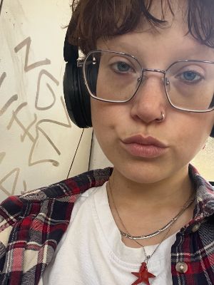 Eine junge Person blickt in die Innenkamera und trägt ein weißes T-Shirt, ein kariertes Hemd sowie Kopfhörer.