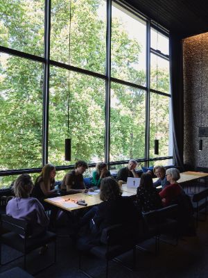 Nine people are sitting together at a long table, chatting. In the background, green leafy trees can be seen through a tall window wall.