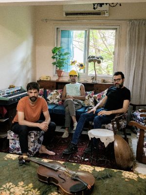 Das Trio sitzt in einem Wohnzimmer voller Instrumente. Eine Person trägt einen Bauhelm.