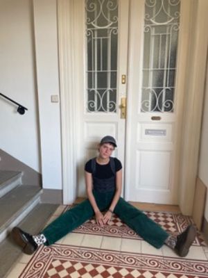Henriette Bayer sitzt vor einer Wohnungstür im Treppenhaus auf dem Boden und sieht direkt in die Kamera.