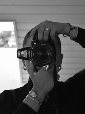 Eine Person steht vor einem Spiegel und macht ein Foto mit der Kamera, die das Gesicht verdeckt. Das Bild ist in Schwarz-Weiß.