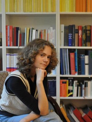 Sophia Becker sitzt vor einem großen Bücherregal und stützt den Kopf auf eine Faust.