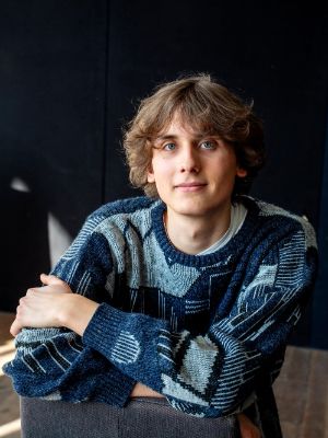 Eine junge Person sitzt auf einem Stuhl vor dunklem Hintergrund und blickt in die Kamera.