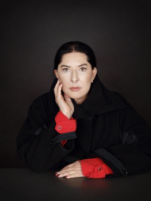 A white woman with black hair and dark eyes looks directly into the camera. She is wearing a black jacket with a red blouse peeking out at the hands. She is sitting at a table, only her upper body is visible. One hand is resting on the table, the other is leaning against her face.