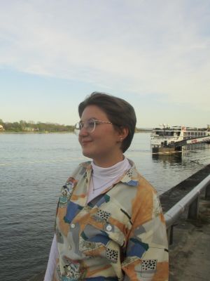 Eine junge Frau steht am Wasser und blickt zur Seite. Sie trägt ein buntes Hemd.