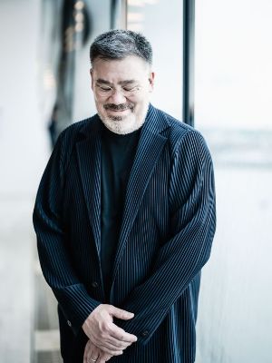 A man in a black coat and jumper stands smiling in front of the camera, looking down with his eyes closed.