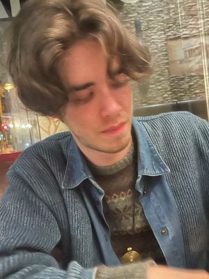 Person mit leichtem Bart und längeren dunkelblonden Haaren sitzt in einem Café und schaut auf den Tisch vor sich.