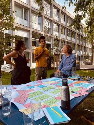 Drei Personen unterhalten sich. Am Tisch neben ihnen ist ein Plakat mit vielen Post-its