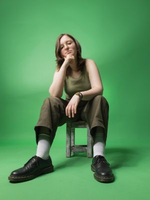 Eine junge Frau sitzt auf einem Hocker vor einem grünen Hintergrund. Das Bild ist von unten fotografiert. Sie stützt ihr Kinn auf ihrem angewinkelten Arm ab und blickt direkt in die Kamera.