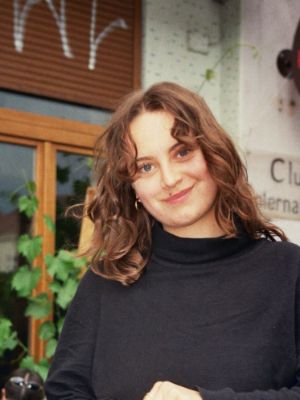 Eine junge Person blickt in die Kamera und lächelt. Sie hat lange braune Haare und trägt einen schwarzen Pullover.