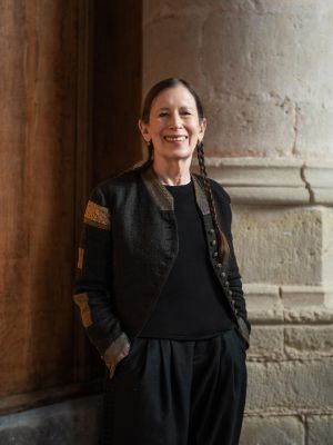 A portrait of Meredith Monk. She stands in front of an old wall and smiles at the camera.
