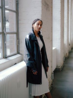 Eine Person in weißem Kleid und schwarzer Lederjacke steht vor einem Fenster.