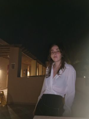 Eine junge Person steht vor einem Haus in dunkler Umgebung und trägt ein weißes Hemd.