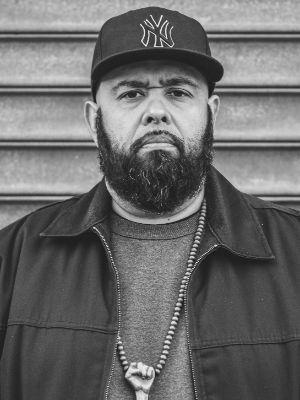 MC Dälek schaut geradeaus in die Kamera. Er trägt einen Vollbart und eine NYY Mütze.