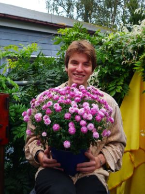 Anton Conrad schaut direkt in die Kamera und hält dabei einen Blumentopf mit violetten Chrysanthemen vor sich.