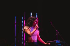 A female read holds a microphone in her hand. Behind her blue and pink stage lights.