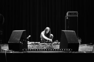 Eine weiblich gelesene Person, die hinter Synthesizern und zwei Lautsprechern auf dem Bühnenboden hockt.  