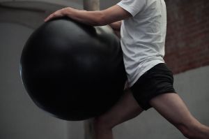 A male read person in a white T-shirt and black shorts holds a black circus ball in front of his body.