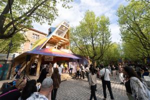 People are standing outside in a square surrounded by trees and a wooden house