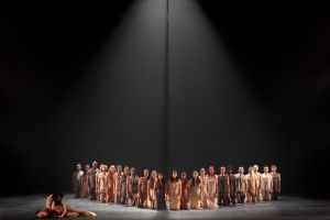 A group of dancers kneel on a stage. A couple is lying on the floor in front of them.