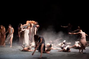 A group of dancers roll around on a gravel-covered stage. One couple carries another couple on their shoulders.