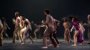 A group of dancers runs around on a gravel-covered stage.