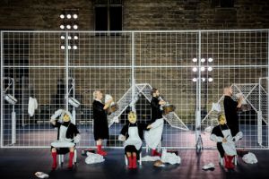Performers with puppet-like masks in front of a barred fence