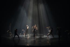 A group of dancers moves in a cone of light on a dark stage.