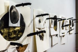 An installation view with various images attached to a wall with black knives.