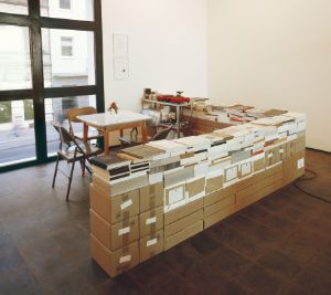 View of Rirkrit Tiravanija's installation "untitled 1993 (café deutschland)", a table with chairs in front of a window. Around the table are boxes with paper catalogues on them.