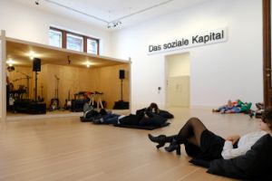Installationsansicht von Rirkrit Tiravanija's "untitled 1996 (rehearsal studio no. 6), 1996" und "untitled 1998 (das soziale kapital) (sign)" mit Besucher*innen im Rahmen der Ausstellung "Rirkrit Tiravanija: DAS GLÜCK IST NICHT IMMER LUSTIG" im Gropius Bau.