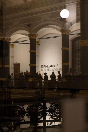 Ein schwarzes Gittersystem mit Schwarz-Weiß-Fotografien der Fotografin Diane Arbus in den Ausstellungsräumen des Gropius Bau. Zwischen den Stellagen sind Besuchende, die sich die Fotografien anschauen.