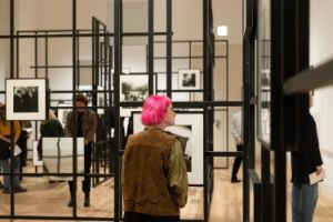 Ein schwarzes Gittersystem mit Schwarz-Weiß-Fotografien der Fotografin Diane Arbus in den Ausstellungsräumen des Gropius Bau. Zwischen den Stellagen sind Besuchende, die sich die Fotografien anschauen.