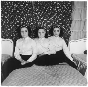 Three female-presenting individuals are seated on a bed. They are triplets, wearing identical clothing and the same hairstyle.